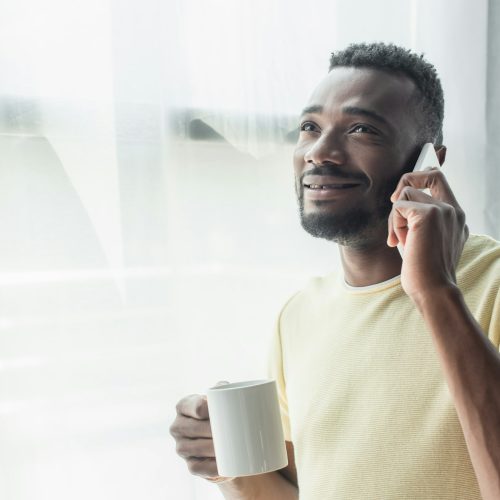 smiling african american man holding mug with tea and talking on cellphone